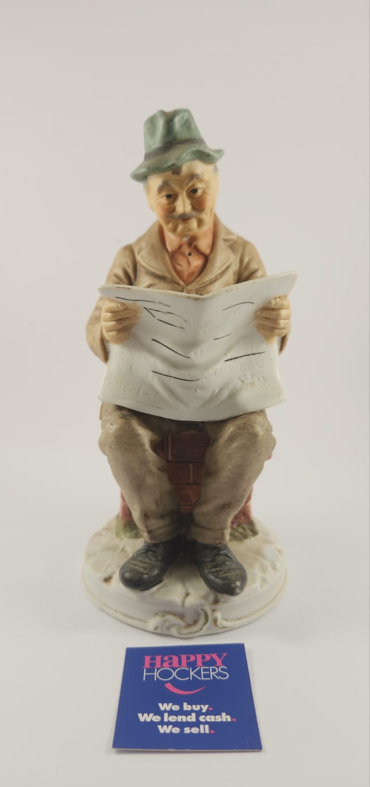 CERAMIC FIGURINE OF MAN SITTING ON BRICK LEDGE READING NEWSPAPER.